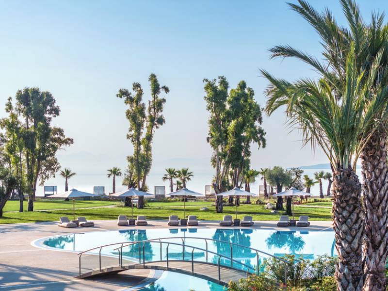 Pool area at Ikos Odisia resort with palm trees, sun loungers, and a view of the beach and sea.
