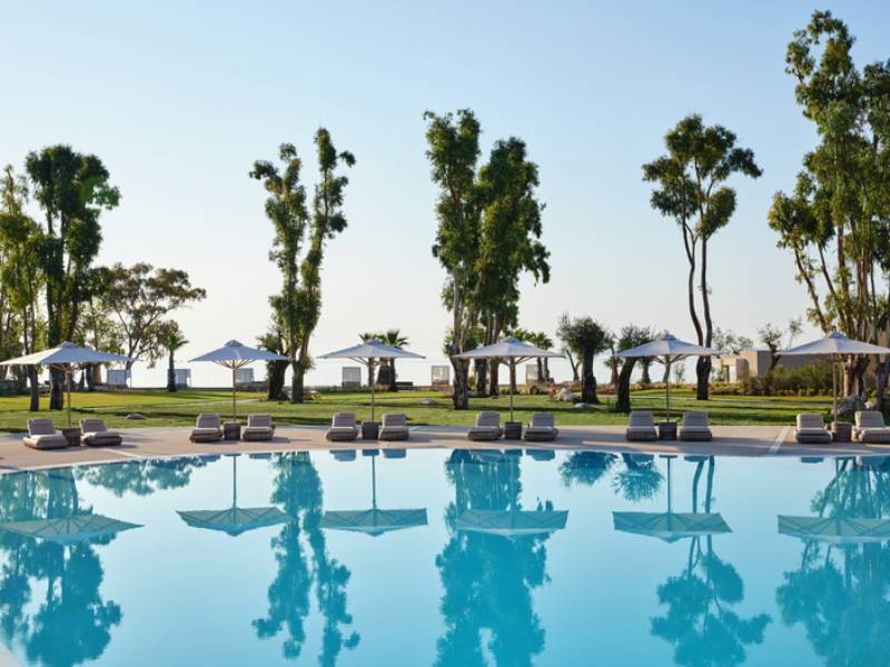 Outdoor pool at Ikos Odisia with surrounding trees, white parasols, and the sea in the background.