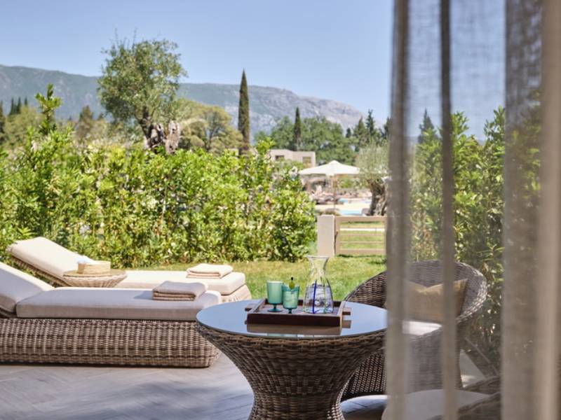Private garden at Ikos Odisia with wicker lounge chair and table, surrounded by greenery and mountain views in the background.
