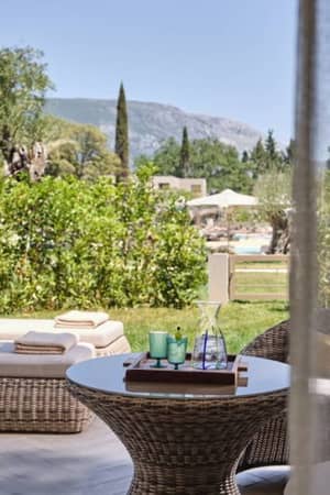 Private garden at Ikos Odisia with wicker lounge chair and table, surrounded by greenery and mountain views in the background.