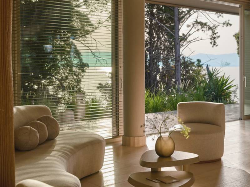 Ikos Odisia spa lounge area with curved seating, a coffee table with flowers, and large windows overlooking greenery and the sea.