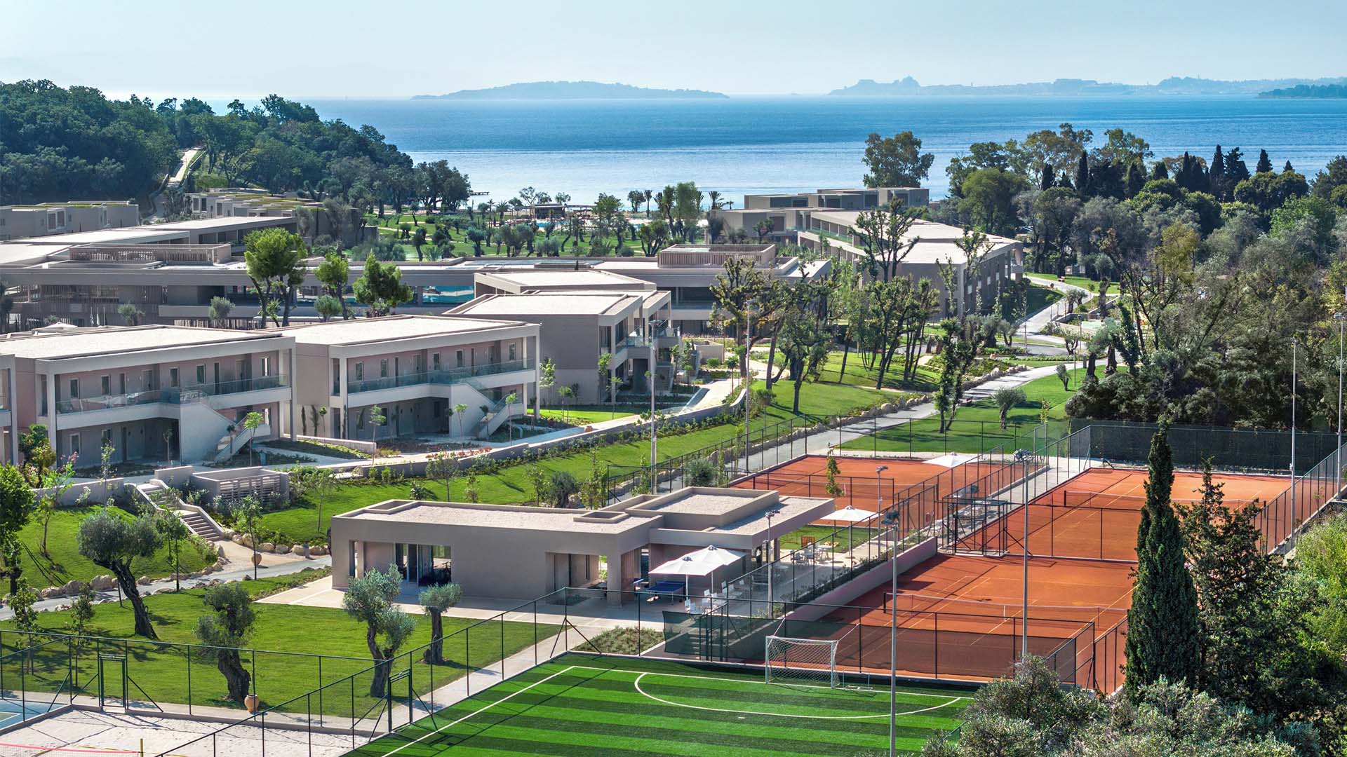 Aerial view of Ikos Odisia resort with modern accommodations, tennis courts, greenery, and a coastal backdrop.