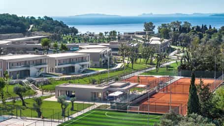 Aerial view of Ikos Odisia resort with modern accommodations, tennis courts, greenery, and a coastal backdrop.