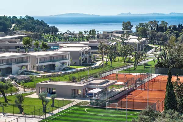 Aerial view of Ikos Odisia resort with modern accommodations, tennis courts, greenery, and a coastal backdrop.