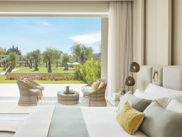 Interior of an Ikos Odisia one-bedroom suite with a private garden view, showcasing a spacious bed, soft pillows, and outdoor seating overlooking a lush landscape.