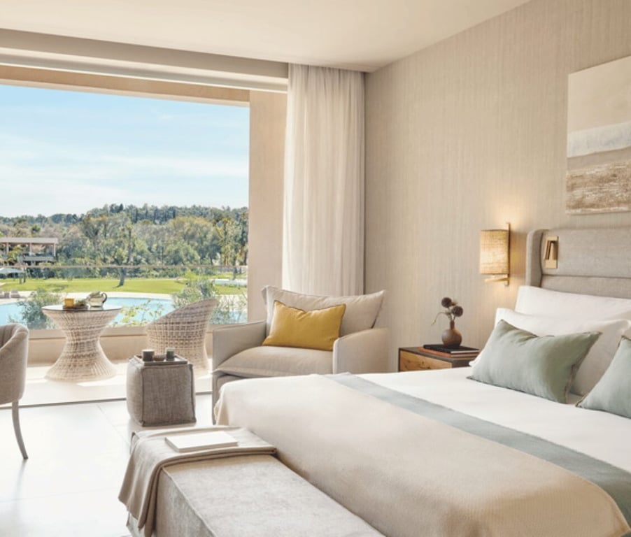 Interior of Ikos Odisia hotel room with pool view, featuring a large bed and a cosy seating area with a backdrop of greenery.
