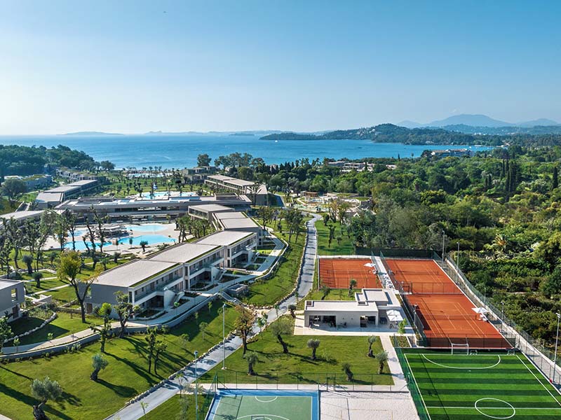 Aerial view of Ikos Odisia resort featuring tennis courts, a pool area, lush gardens, and the Ionian Sea.