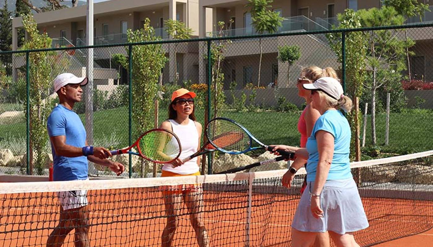 Adults playing tennis at Ikos Odisia Resort on a clay court, surrounded by modern buildings and greenery, exchanging greetings over the net.