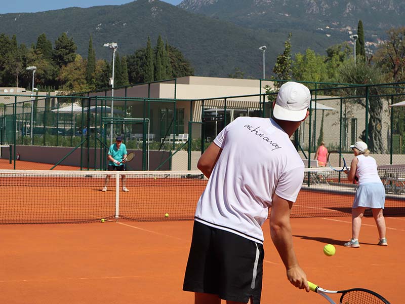 Tennis players on a court with a man in a white shirt and cap preparing to hit the ball, set against a mountainous backdrop.