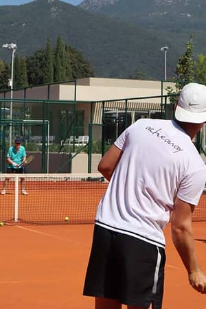 Tennis players on a court with a man in a white shirt and cap preparing to hit the ball, set against a mountainous backdrop.