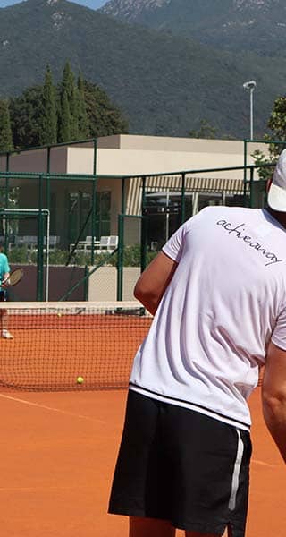 Tennis players on a court with a man in a white shirt and cap preparing to hit the ball, set against a mountainous backdrop.