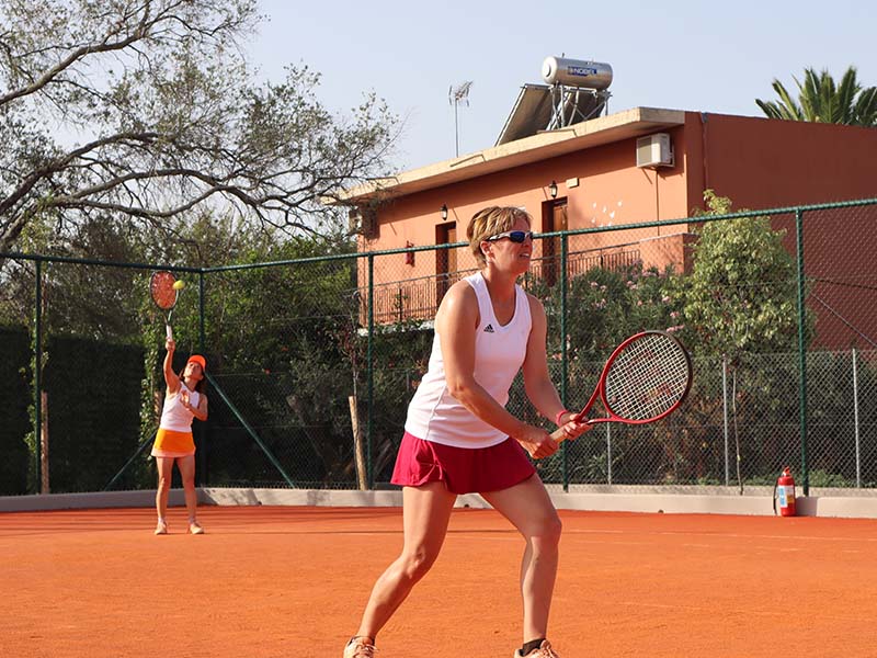 Woman in red shorts ready to volley on a clay tennis court, with an opponent serving in yellow skirt, surrounded by a fenced area with trees and a building with solar panels.