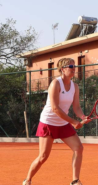 Woman in red shorts ready to volley on a clay tennis court, with an opponent serving in yellow skirt, surrounded by a fenced area with trees and a building with solar panels.