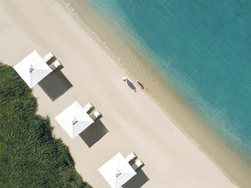 Aerial view of Ikos Olivia beach, showing three white sun loungers with umbrellas and two people walking by the turquoise sea.