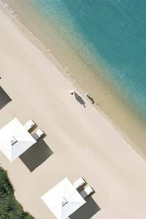 Aerial view of Ikos Olivia beach, showing three white sun loungers with umbrellas and two people walking by the turquoise sea.