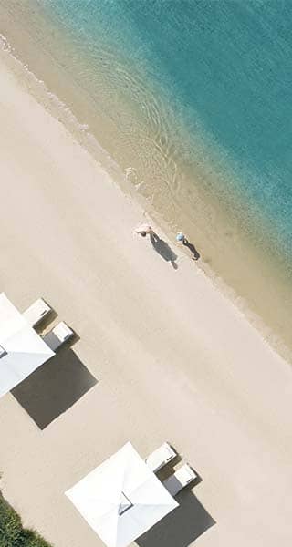 Aerial view of Ikos Olivia beach, showing three white sun loungers with umbrellas and two people walking by the turquoise sea.