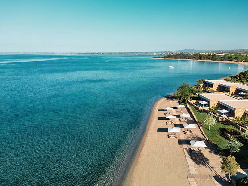Aerial view of Ikos Olivia Resort's beach with sun loungers, parasols, and clear Aegean Sea waters.
