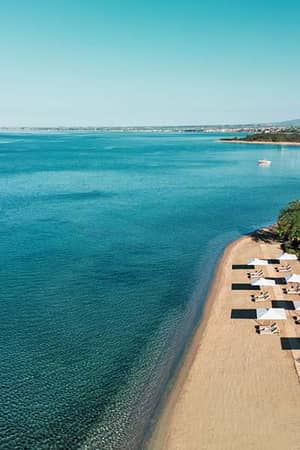 Aerial view of Ikos Olivia Resort's beach with sun loungers, parasols, and clear Aegean Sea waters.