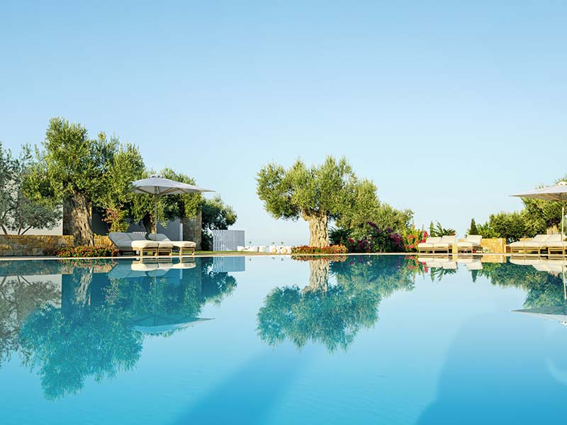 Tranquil swimming pool at Ikos Olivia with sun loungers, parasols, and olive trees reflecting in the blue water.