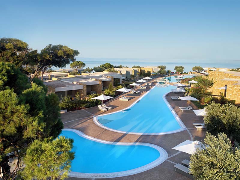 Aerial view of Ikos Olivia resort with a winding swimming pool, lush gardens, and the Aegean Sea in the background.