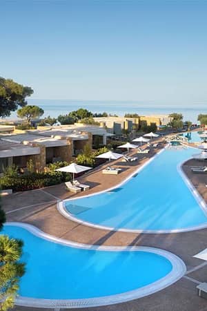 Aerial view of Ikos Olivia resort with a winding swimming pool, lush gardens, and the Aegean Sea in the background.