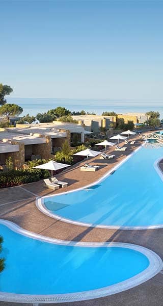 Aerial view of Ikos Olivia resort with a winding swimming pool, lush gardens, and the Aegean Sea in the background.