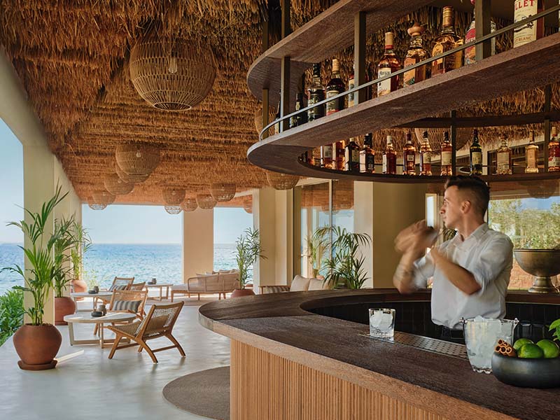 Beachfront bar at Ikos Olivia resort with a mixologist, sea view, thatched roof, and elegant seating.