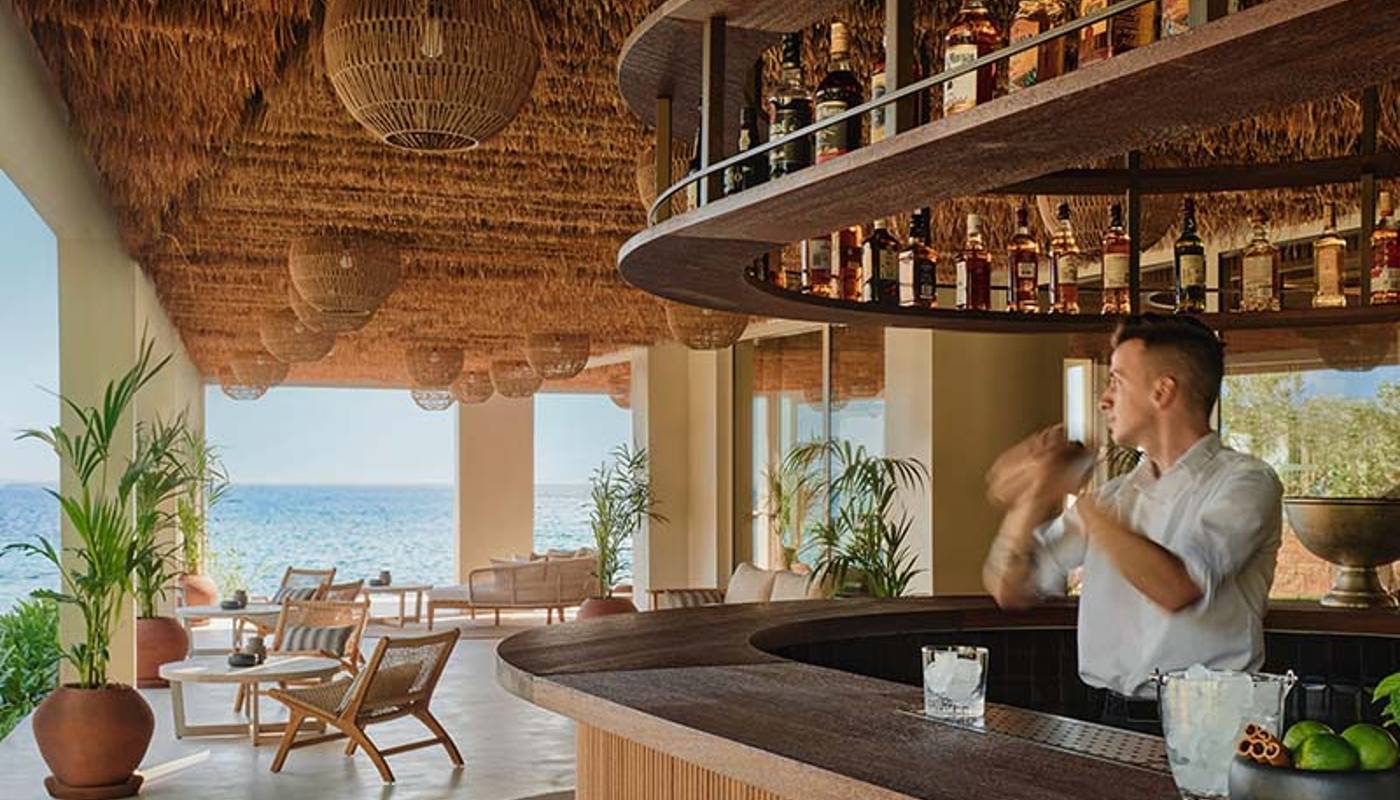 Beachfront bar at Ikos Olivia resort with a mixologist, sea view, thatched roof, and elegant seating.