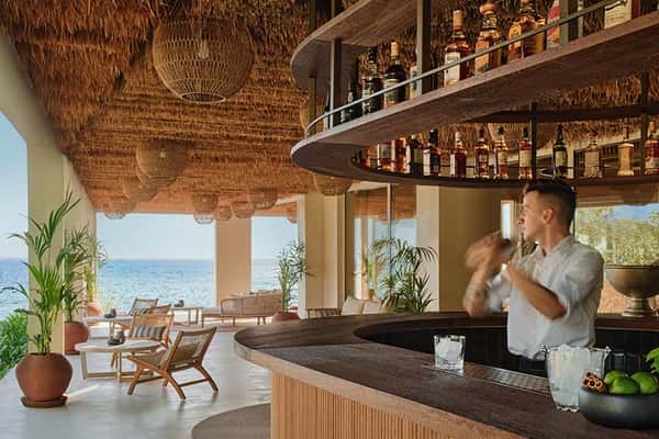Beachfront bar at Ikos Olivia resort with a mixologist, sea view, thatched roof, and elegant seating.
