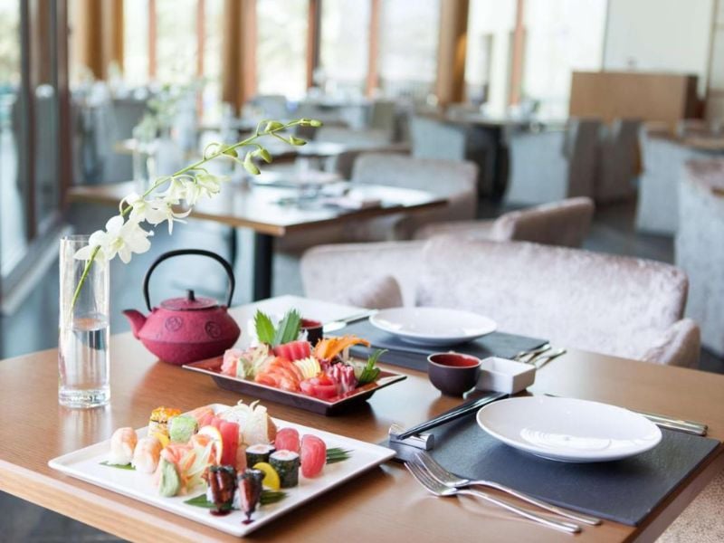 Table set with sushi and sashimi at JA Gallery, including a red teapot and white orchid flowers, in a modern dining room with elegant furnishings.