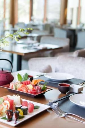 Table set with sushi and sashimi at JA Gallery, including a red teapot and white orchid flowers, in a modern dining room with elegant furnishings.