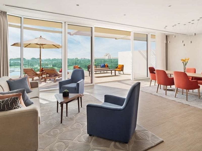 Modern living room with large glass doors, leading to a patio with loungers and a landscape view, featuring plush sofas, armchairs, and a dining table with red chairs.
