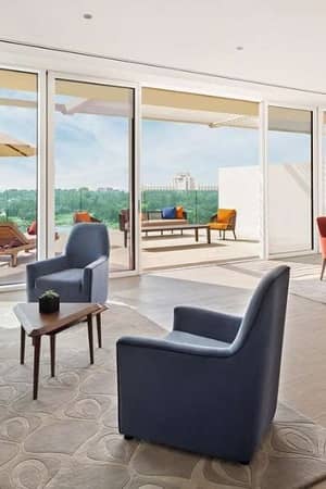 Modern living room with large glass doors, leading to a patio with loungers and a landscape view, featuring plush sofas, armchairs, and a dining table with red chairs.