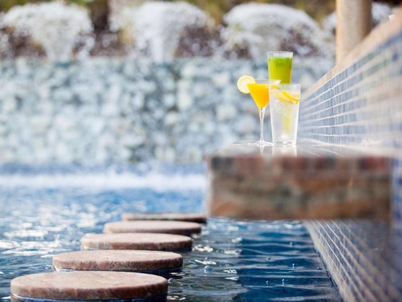 Three colourful drinks on a ledge at a pool bar with cascading water features in the background, creating a relaxing environment.