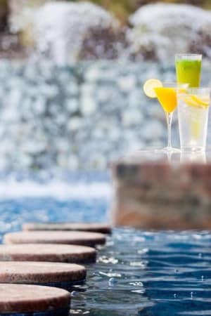 Three colourful drinks on a ledge at a pool bar with cascading water features in the background, creating a relaxing environment.