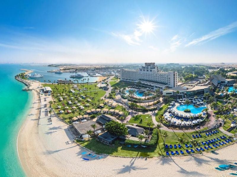 Aerial view of JA The Resort with pools, gardens, and a beachfront along turquoise waters under a sunny sky.