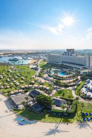 Aerial view of JA The Resort with pools, gardens, and a beachfront along turquoise waters under a sunny sky.