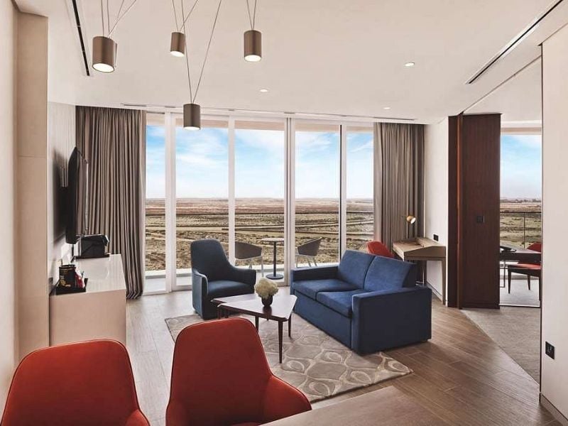 Modern hotel room with blue sofa, red chairs, large TV, and floor-to-ceiling windows overlooking a desert landscape.