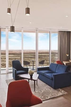 Modern hotel room with blue sofa, red chairs, large TV, and floor-to-ceiling windows overlooking a desert landscape.