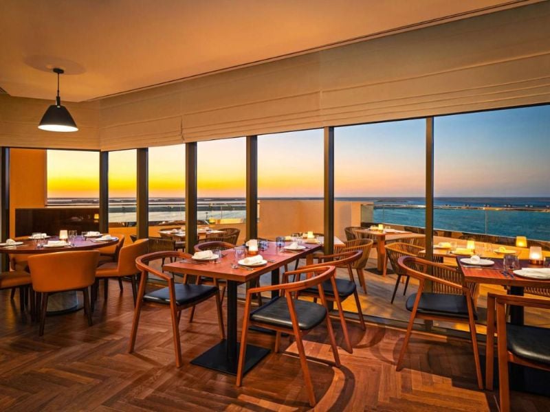 Restaurant interior with tables and chairs, large windows displaying a scenic ocean sunset view, creating a warm and inviting dining atmosphere.
