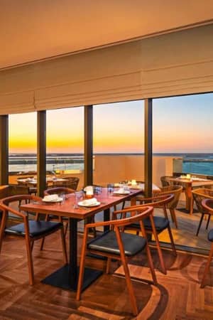 Restaurant interior with tables and chairs, large windows displaying a scenic ocean sunset view, creating a warm and inviting dining atmosphere.