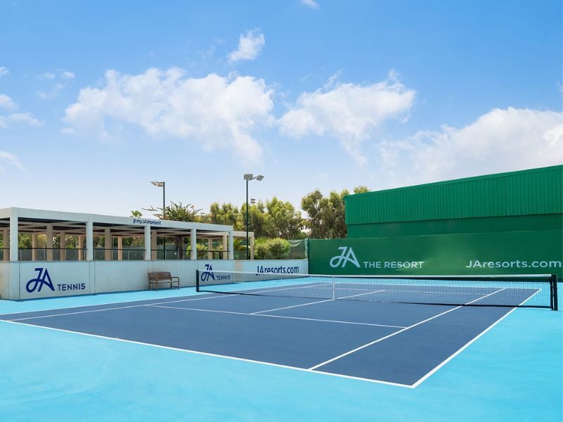 Outdoor tennis court at JA Gallery with blue playing surface, green surroundings, and JA Resorts branding.