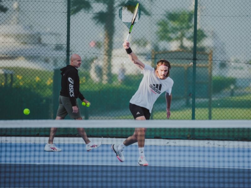 Male tennis player in a white Adidas outfit executing a forehand swing on an outdoor court with a coach in the background.