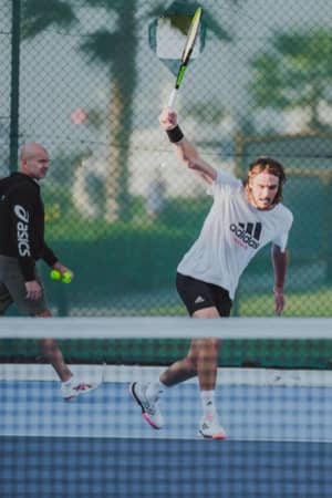 Male tennis player in a white Adidas outfit executing a forehand swing on an outdoor court with a coach in the background.