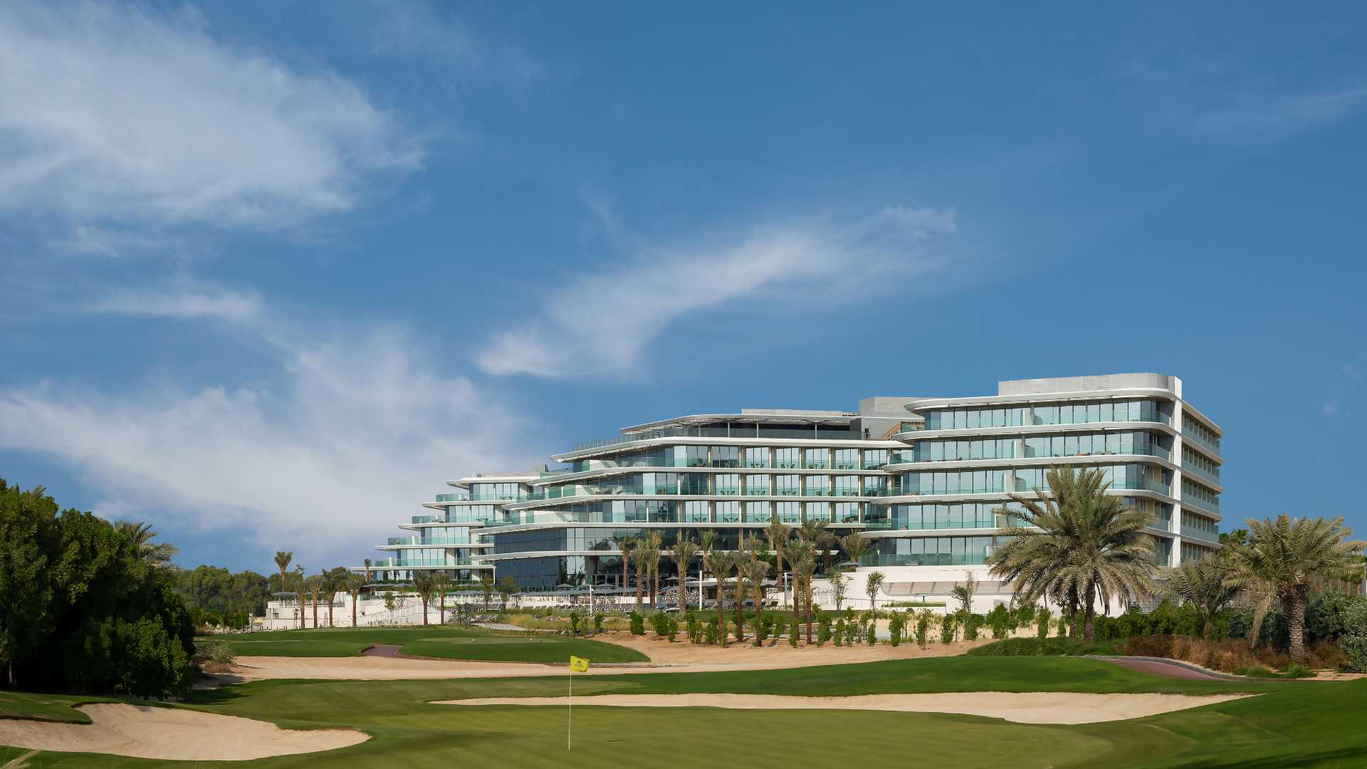 Panoramic view of JA Lakeview Hotel with modern glass façade, surrounded by a golf course and palm trees under a blue sky.