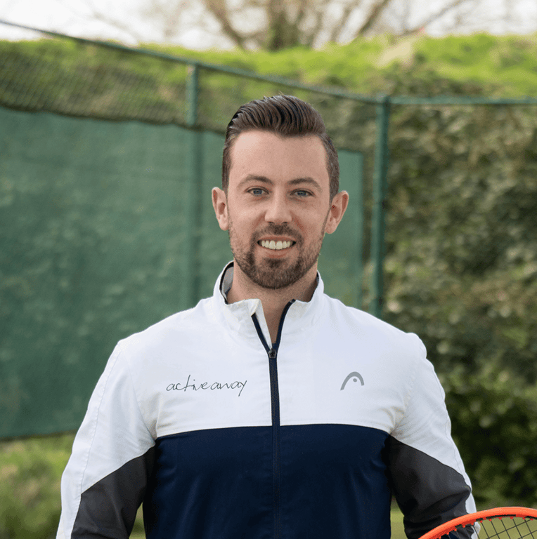 Joshua Thompson, MD of Active Away, stands on a tennis court wearing a branded jacket and holding a tennis racket.