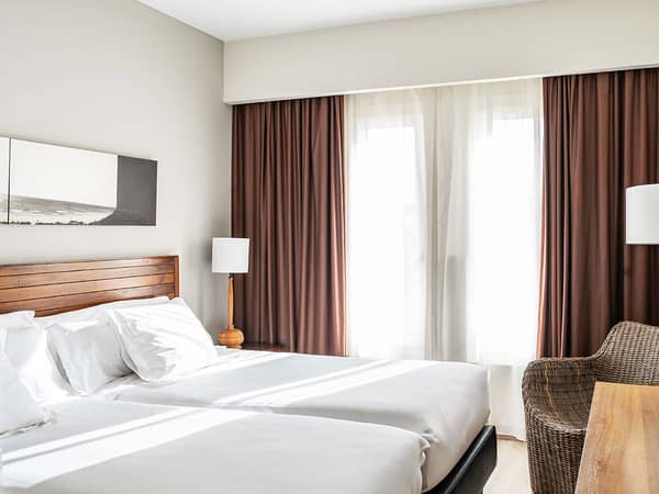 Junior Suite at Hotel Vincci with twin beds, white linens, wooden headboards, and large windows with brown drapes.