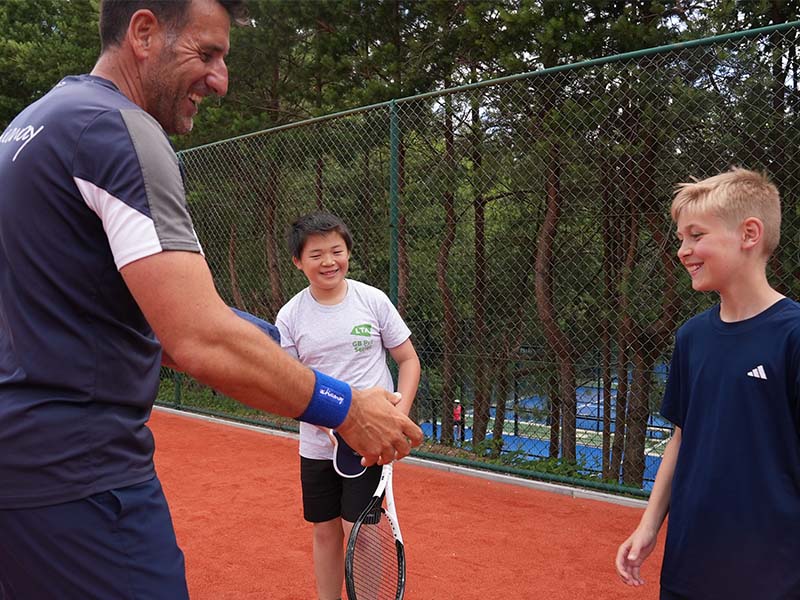 A tennis coach speaking with two young boys on a clay court surrounded by trees at The Lensbury junior tennis camp.