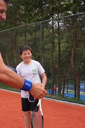A tennis coach speaking with two young boys on a clay court surrounded by trees at The Lensbury junior tennis camp.