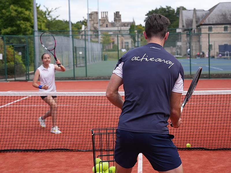 A coach assisting a young girl during a tennis practice session on a red clay court at The Lensbury, with a historic building in the background.